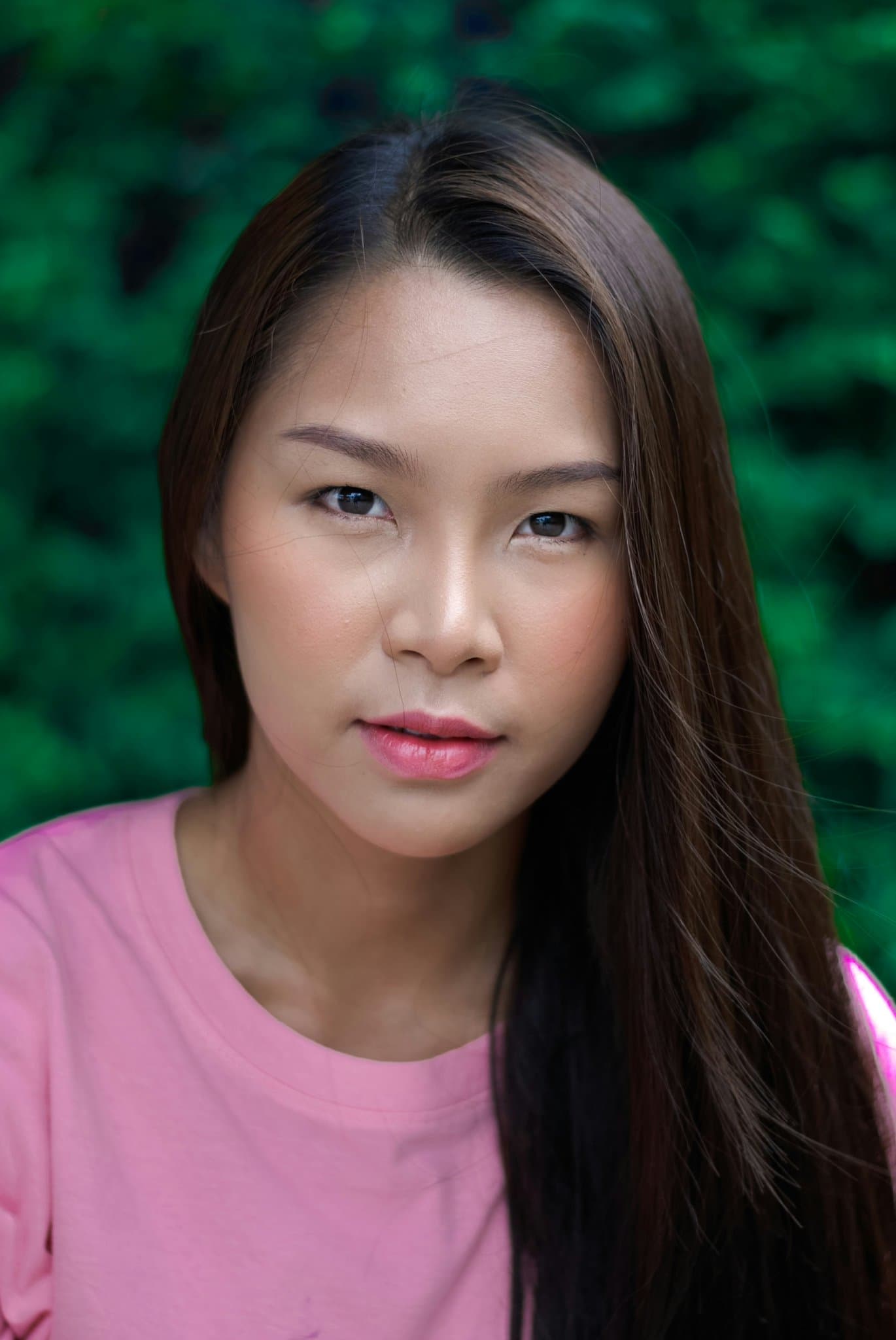 Portrait with natural makeup, pink top, long straight hair, green blurred background.