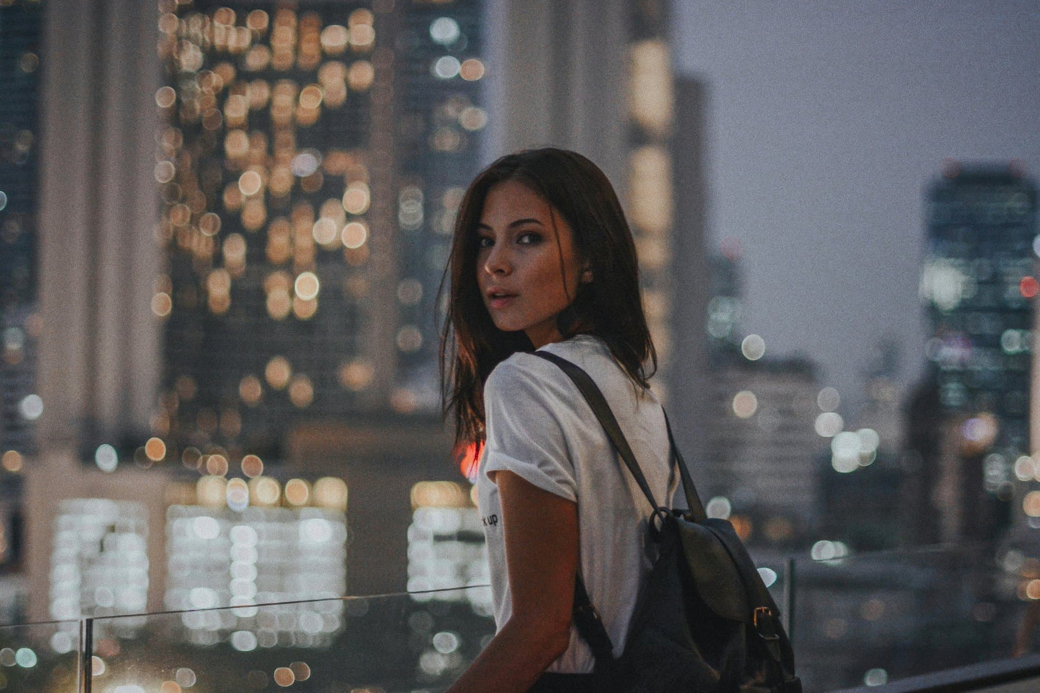 City rooftop scene with soft bokeh lights framing a backpacked figure turning toward the camera.